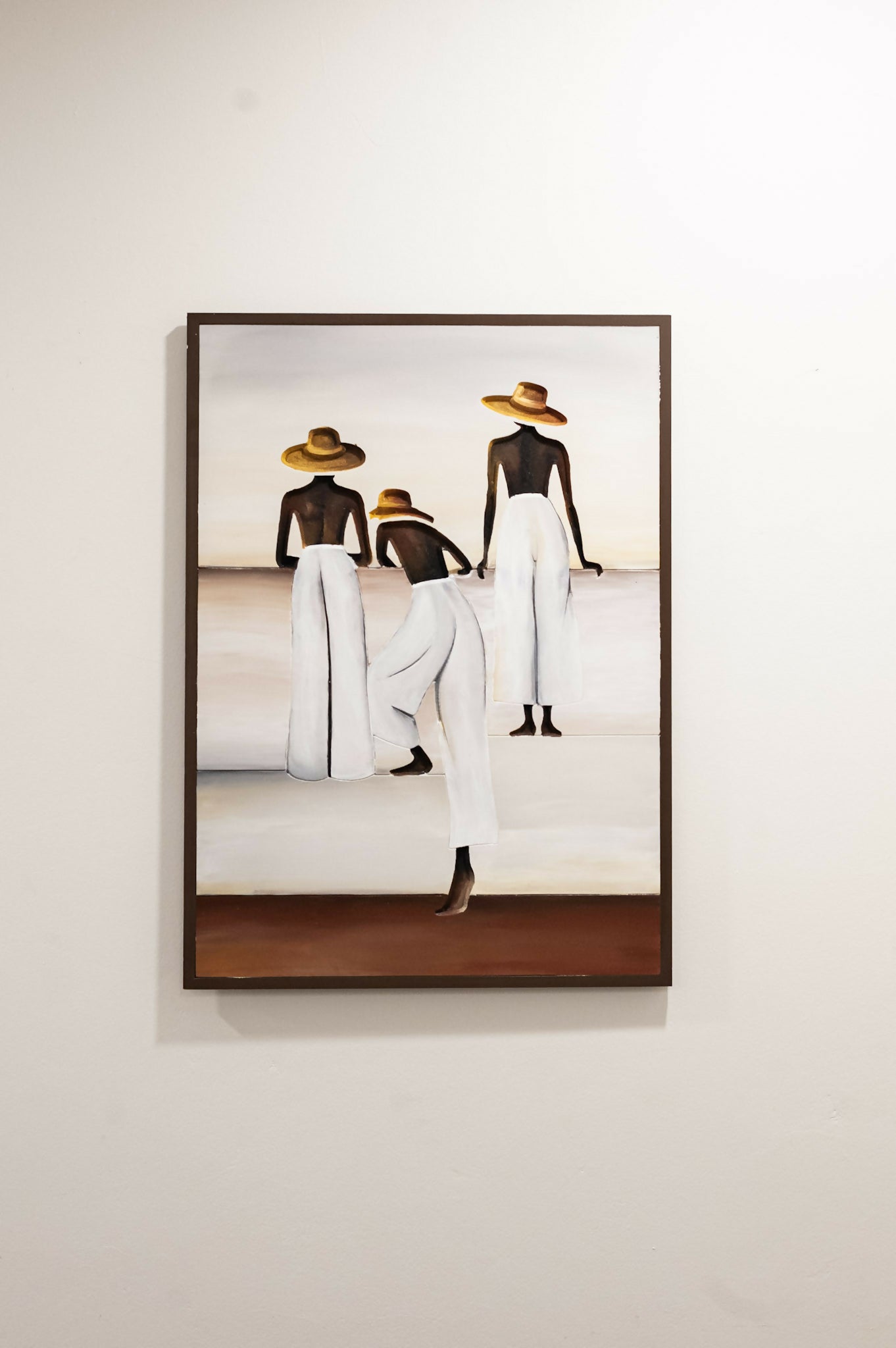 A framed painting on a light beige wall, depicting three stylized figures from behind wearing white garments and straw hats, with a dark brown frame. Caribbean Art