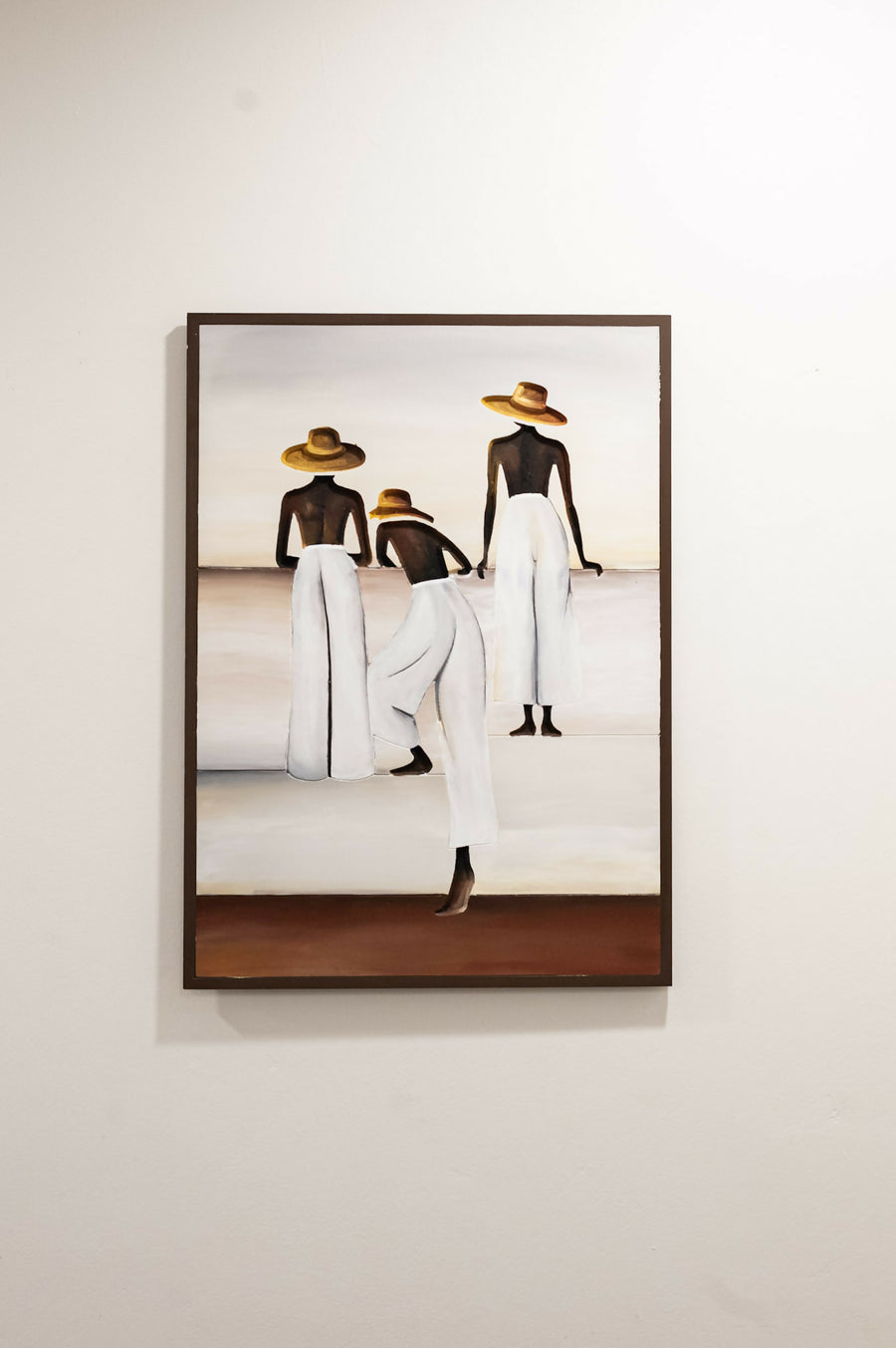 A framed painting on a light beige wall, depicting three stylized figures from behind wearing white garments and straw hats, with a dark brown frame. Caribbean Art