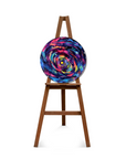 A round canvas print featuring a vibrant, swirling galaxy design in shades of blue, purple, pink, and yellow, displayed on a wooden easel against a solid white background.