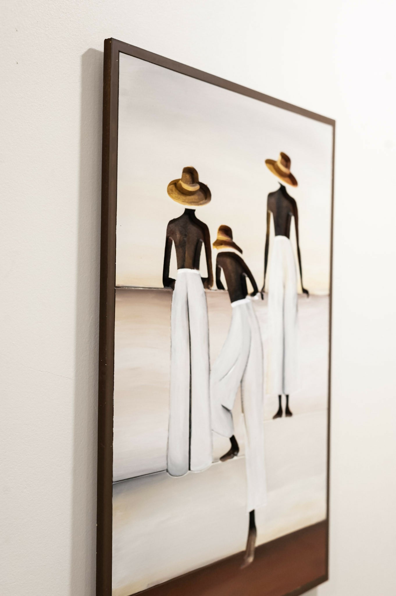 A framed art print depicting three fashion figures from behind wearing white pants, black tops, and wide-brimmed straw hats, hanging on a light beige wall. Caribbean Art 